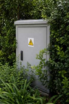 A gray electrical box surrounded by dense bushes with a warning sticker displayed prominently.