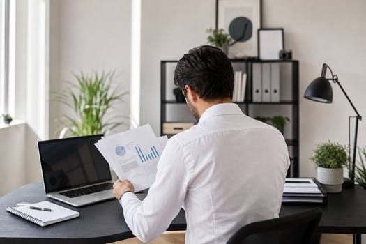 Back view focused ethnic male in white shirt reading document while sitting at desk with netbook in light office